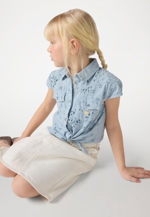 Light blue short-sleeve shirt with eyelet pattern and a tied front, paired with a white knee-length skirt. Sitting pose, neutral background.