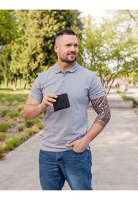 Gray polo shirt with short sleeves, blue jeans, tattooed arm, holding a black wallet, in a park setting with greenery in the background.