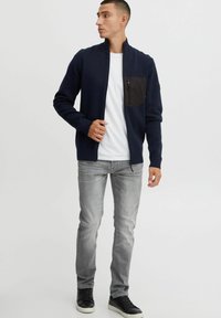 Chaqueta de navy con cremallera y un bolsillo negro en el pecho, textura de punto, combinada con jeans grises y zapatillas negras. Diseño casual y ajustado.