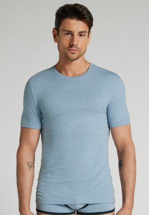 Junger Mann mit kurzen dunklen Haaren, der ein eng anliegendes hellblaues kurzärmliges Hemd und passende Unterwäsche trägt und vor einem einfarbigen Hintergrund steht.