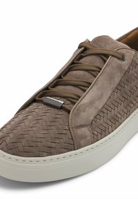 Brązowe zamszowe sneakersy z plecioną teksturą, okrągłym noskiem i kontrastową, kremowo-białą gumową podeszwą. Posiadają płaskie sznurowadła i metalową tabliczkę z logo.