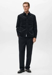 Camicia in corda nera con due tasche sul petto, chiusura con bottoni e maniche lunghe, abbinata a pantaloni scuri e scarpe nere.