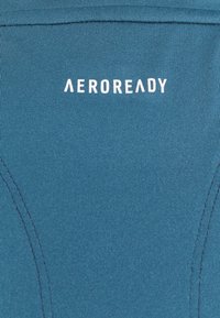 Haut de sport AEROREADY en sarcelle, avec une texture côtelée, des coutures plates et un logo blanc bien visible. Le tissu est anti-transpiration et extensible.