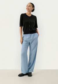 Schwarze Kurzarm-Cardigan mit Knöpfen, kombiniert mit hellblauen, weit geschnittenen Hosen. Schwarze Schuhe runden den Look ab, mit subtiler Textur und tailoring.