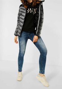 Svart och grå kviltad pufferjacka, svart hoodie med grafisk text, blå blekta slim jeans, krämfärgade sneakers, står mot en ljus bakgrund.