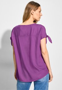 Blusa viola con vestibilità rilassata, maniche corte e dettagli con fiocchi sui lati. Presenta un orlo arrotondato e una consistenza liscia.
