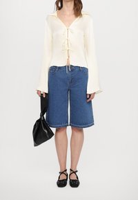 Cremefarvet langærmet top med V-hals og bindedetalje foran, kombineret med blå denim Bermudashorts. Sort skuldertaske og flade sko med stropper.