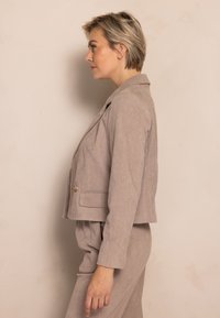 Hellbraune Cord-Feinstrickjacke mit figurbetontem Schnitt, Revers mit Kerbkragen, Vordertaschen und Knopfverschluss, kombiniert mit passenden Hosen.