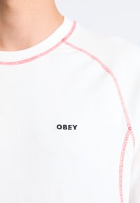 White cotton t-shirt with round neckline, red stitching accents, and "OBEY" logo in black embroidered on the chest.