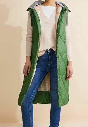 Osoba ubrana w zieloną pikowaną kamizelkę z kapturem, beżowy sweter z dekoltem w serek i niebieskie jeansy na jednolitym beżowym tle.