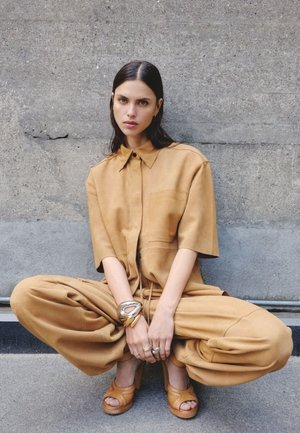 Világosbarna, rövid ujjú, bő szabású ing, elülső zsebbel, és hozzá illő, laza nadrággal; világosbarna platform szandállal és rétegezett fém karkötőkkel párosítva.