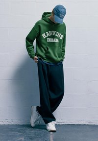 Zelená mikina s kapucňou s bielym potlačeným nápisom "HAWKINS INDIANA", v kombinácii s voľnými čiernymi nohavicami a bielymi teniskami. Modrá džínsová čiapka s textovým detailom.