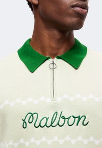 Personne portant une chemise de couleur claire avec un col vert et une fermeture éclair, brodée du mot "Malbon" en fil vert.
