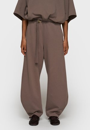 Osoba nosząca luźne spodnie w kolorze taupe o szerokich nogawkach z pasującym paskiem i czarne trampki, stojąca na tle jednolitego tła.