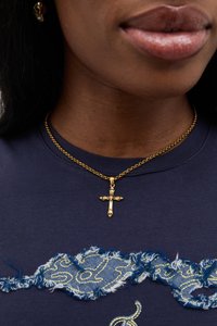 En gullkorsanheng på en kjedehalskjede hviler på en mørkeblå, teksturert crew-neck skjorte med slitne denimdetaljer.