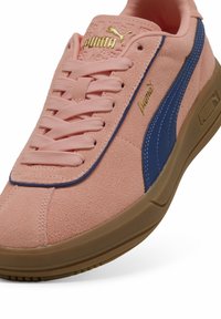 Zapatillas Puma con upper de ante rosa, acentos de cuero azul, logotipo dorado y suela de goma beige con detalles texturizados.