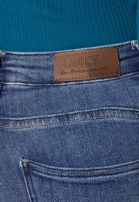Jeans din denim de culoare albastru mediu, cu o suprafață texturată. Prezintă o etichetă din piele maro inscripționată "ONLY" și un buzunar din spate cu cusături curate.
