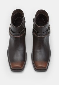 Botas de tobillo de cuero en marrón oscuro, con puntera cuadrada, textura suave, cremalleras laterales y una correa con acentos metálicos.