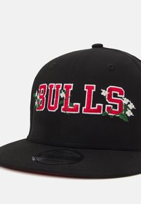 Gorra negra con visera plana, con "BULLS" bordado en rojo y flores blancas y enredaderas verdes como acentos. Tejido texturizado.