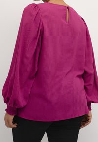 Femme portant un blouse magenta vif avec des manches longues bouffantes et une petite fermeture à bouton au dos du cou, associée à un pantalon noir.