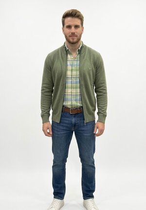 Joven de pie contra fondo blanco que lleva un suéter verde con cremallera, camisa de cuadros, vaqueros azules, cinturón marrón y zapatillas blancas.