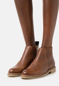 Botas de tobillo de cuero marrón con un acabado texturizado, cremallera lateral y una suela de color claro. Diseño minimalista sin hardware adicional ni patrones.
