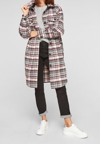 Karierte Jacke in Grau, Weiß und Burgund mit zwei Brusttaschen. Kombiniert mit einem grauen Pullover, schwarzen hochgekrempelten Hosen und weißen Turnschuhen.