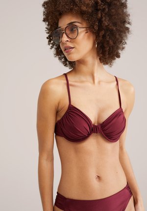 Donna con capelli ricci che indossa occhiali da sole rotondi e un bikini bordeaux con dettagli di tessuto arricciato su uno sfondo semplice.