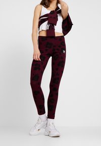 Leggings burdeos con un patrón floral negro, hechos de una tela elástica, combinados con un top corto de canalé blanco y zapatillas blancas.