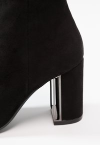 Black suede ankle boot featuring a square toe, a chunky heel with a metallic accent, and a smooth texture on the upper surface.