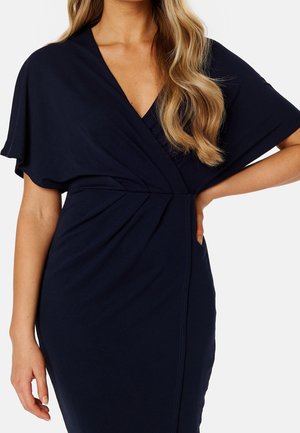 Vrouw in een donker marineblauwe wikkeljurk met korte zwierige mouwen en los blond haar, hand rustend op de heup.