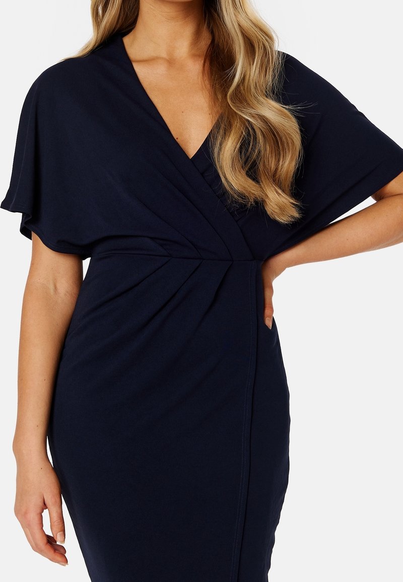Femme en robe portefeuille bleu marine foncé avec de courtes manches volantées et cheveux blonds lâchés, main posée sur la hanche.