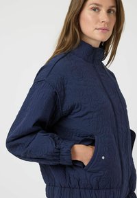 Marinblå vadderad jacka med blommönster, hög krage, elastiska ärmslut och elastisk midja. Har sidofickor med tryckknappsstängning.