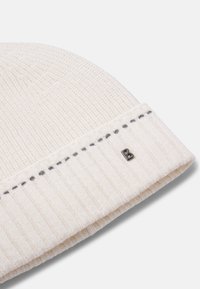 Biała dzianinowa czapka z prążkowaną fakturą, z podwiniętym mankietem i małym metalicznym logo po boku. Subtelne szare detale szwów.