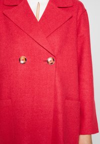 Abrigo de mezcla de lana roja con frente de doble botonadura, cuello con muescas, dos grandes bolsillos y botones redondos con centros contrastantes.