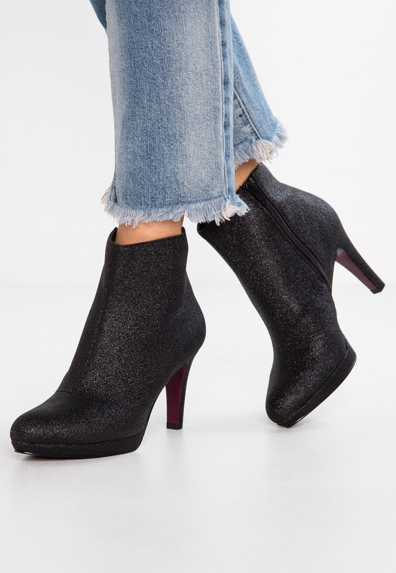 Bottes noires scintillantes à talons hauts et zip latéral portées avec un jean bleu clair à ourlet effiloché sur un sol blanc.
