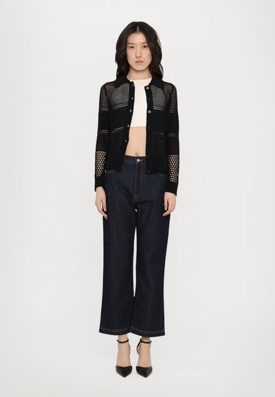 Cardigan nero a maglia con design a rete, abbinato a una crop top bianca e jeans a gamba larga blu scuro. Presenta tacchi a punta neri.