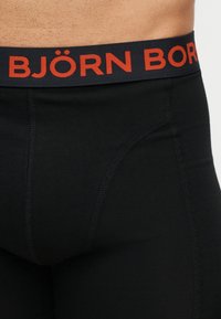 Κοντινό πλάνο μαύρων μποξεράκια με κόκκινο λογότυπο "BJÖRN BORG" στη μέση, που φοριούνται στη μέση και το πάνω μέρος του μηρού ενός ατόμου.