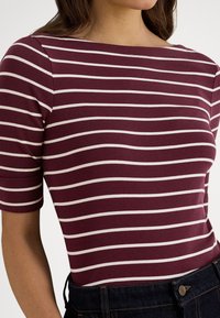 Žena oblečená v maroonovom topu s bielymi horizontálnymi pruhmi a rukávmi do lakťa, zapnutá do tmavomodrých džínsových nohavíc.
