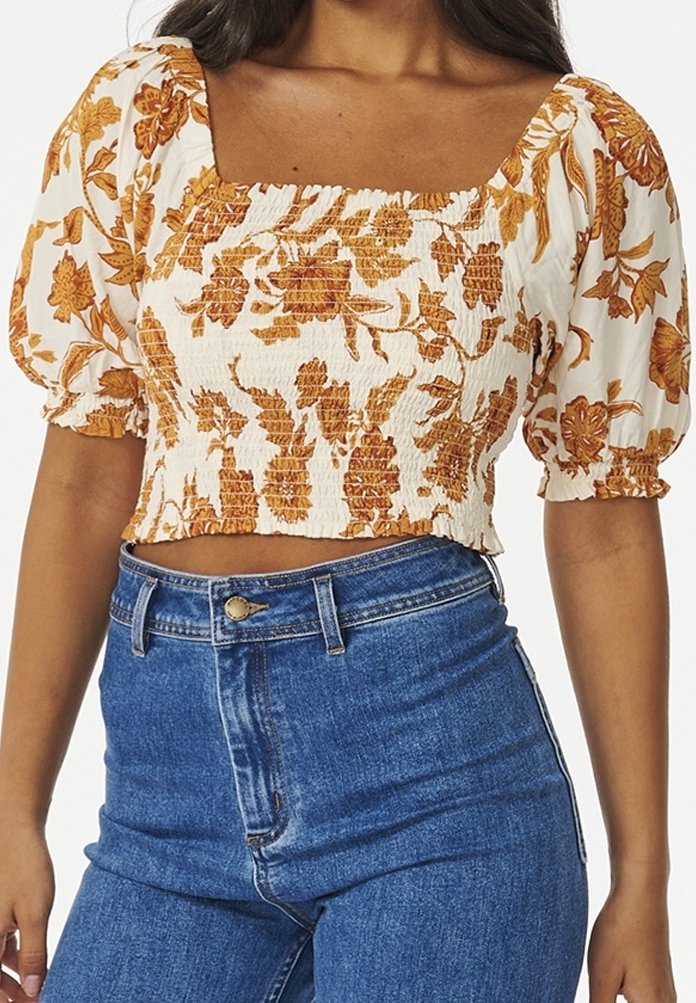 Femme portant un haut blanc smocké court avec un imprimé floral orange et des manches courtes bouffantes, associé à un jean taille haute bleu.