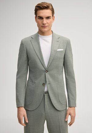 Giovane uomo che indossa una giacca da completo grigio chiaro su misura e pantaloni abbinati sopra una t-shirt bianca, in piedi contro uno sfondo semplice.