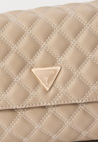 Beige quilted synthetic leather handbag with white stitching and a triangular metallic logo plate featuring "GUESS" and a question mark.