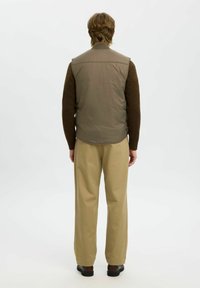 Khakifarbene wattierte Weste mit hohem Kragen, getragen über einem braunen Langarmshirt, kombiniert mit beigen Hosen und schwarzen Schuhen. Neutrale Farbpalette.