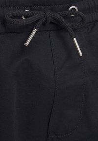 Pantalon en tissu noir avec un cordon de serrage noir noué, doté d'embouts métalliques argentés et d'une poche cousue visible sur le côté droit.