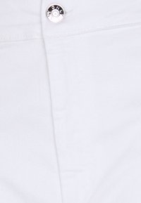 Gros plan sur un tissu blanc avec un bouton en métal et des coutures visibles, probablement une partie d’un vêtement comme un pantalon ou une jupe.