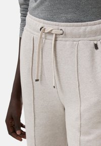 Helle beige Jogginghose mit einem Kordelzug-Bund, weicher Textur und Seitentaschen. Dezenter Glanz auf dem Stoff, mit dunklen Akzenten.