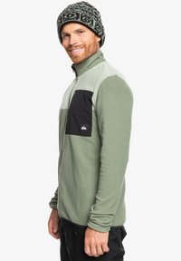 Kurtka polarowa w jasnym zielonym kolorze z czarną kieszenią na zamek, pełnym zamkiem błyskawicznym i długimi rękawami. Model nosi wzorzystą dzianinową czapkę.