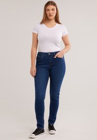 Tânără purtând un tricou alb cu mâneci scurte, blugi skinny de culoare albastru închis și adidași negri, stând cu o mână în buzunar.