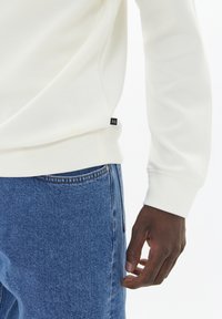 Balta sudraba jaka ar garām piedurknēm un ribotu aproci, apvienota ar ziliem džinsiem. Ietver mazu melnu birku pie apakšmalas.