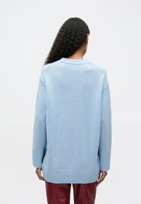 Person mit langen lockigen Haaren, der einen hellblauen oversized Pullover und rote Hosen trägt, mit dem Rücken zu einem schlichten weißen Hintergrund.}}